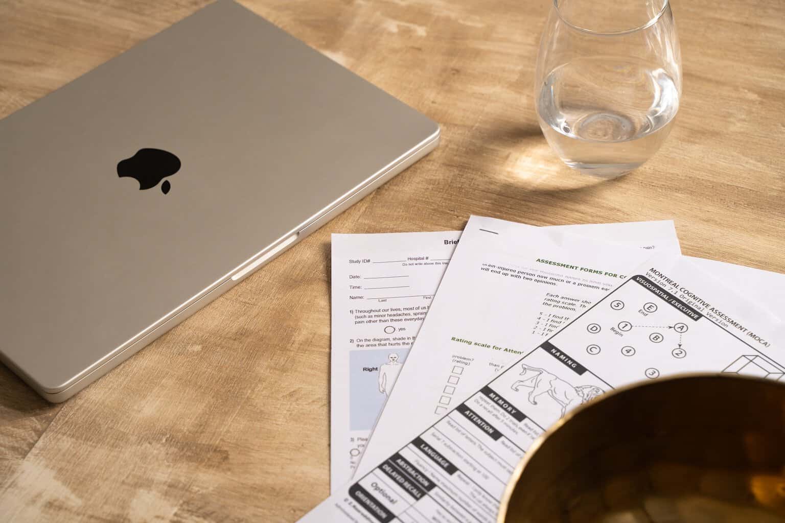 Independent OT Services: Laptop, water glass, and cognitive assessment paperwork on a wooden surface. Illustrates occupational therapy work and cognitive evaluation.